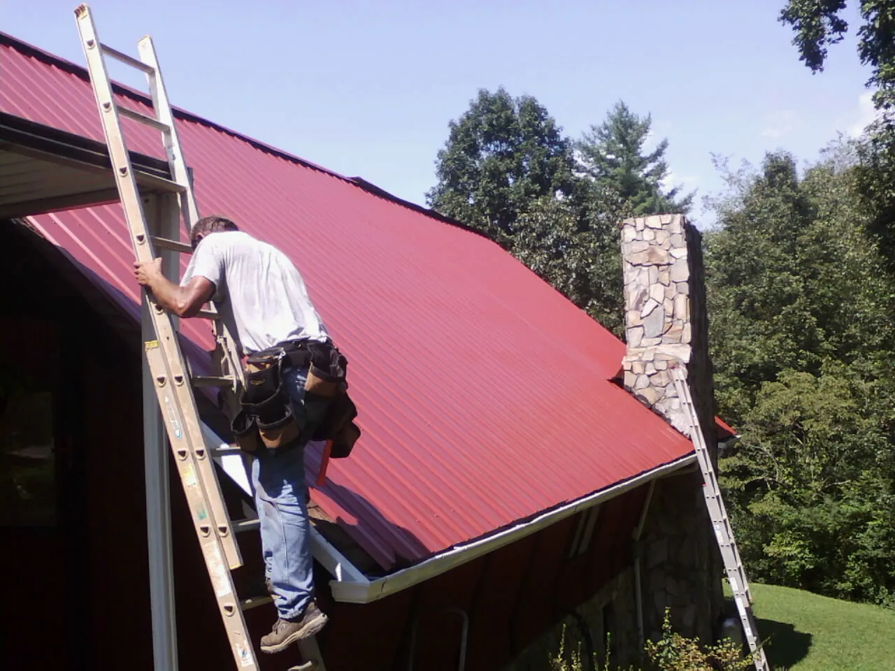 Roofer on a ladder beside a red metal roof and stone chimney — Soffit & Fascia Repair by Roofing Brothers in Hoffman
